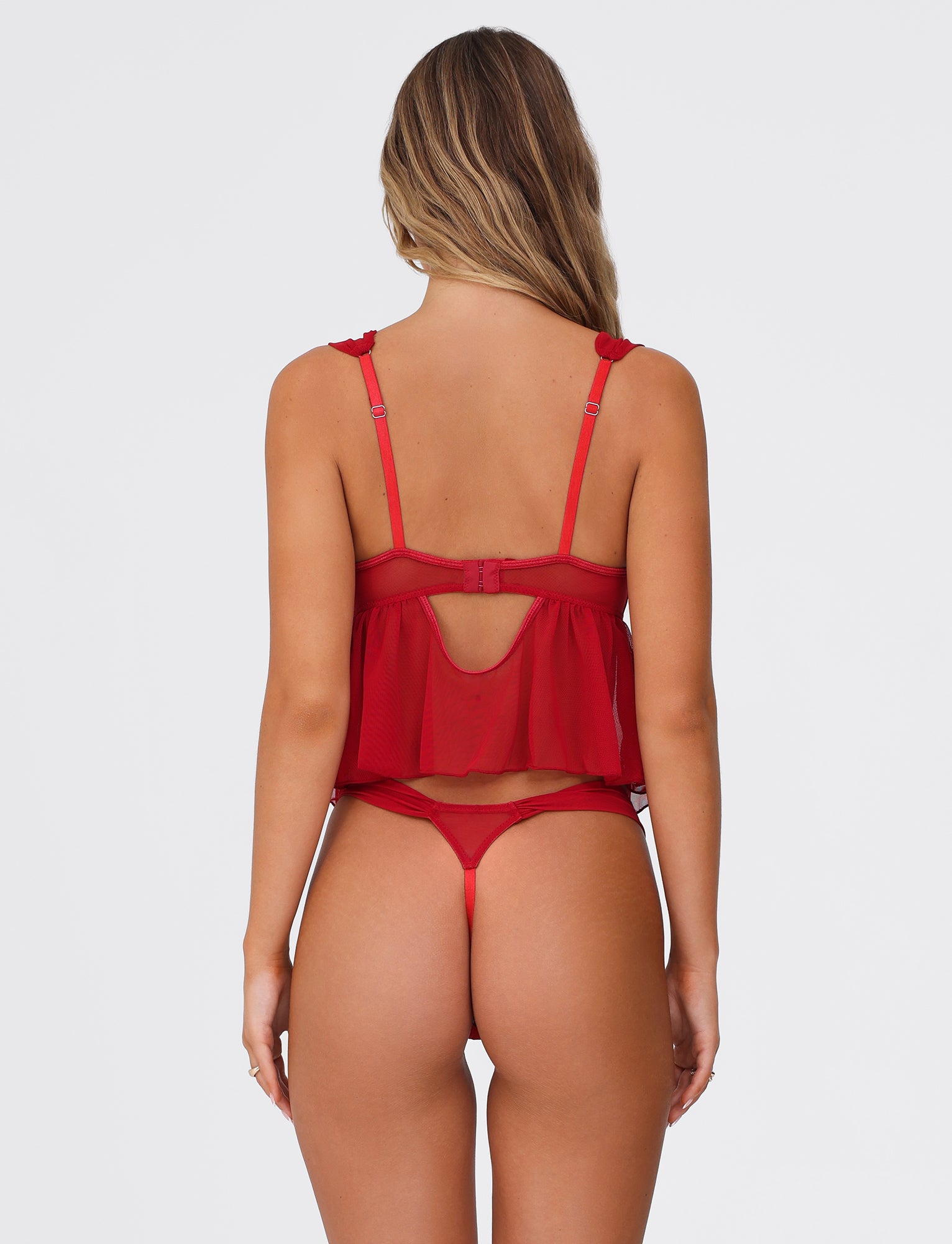A person with long wavy hair is shown from the back wearing the LUANA SET - RED by TIGERMIST, which consists of a crop top with adjustable shoulder straps and a keyhole cutout, paired with matching thong underwear. The babydoll-inspired set features stretch lace detail. The background is plain white.