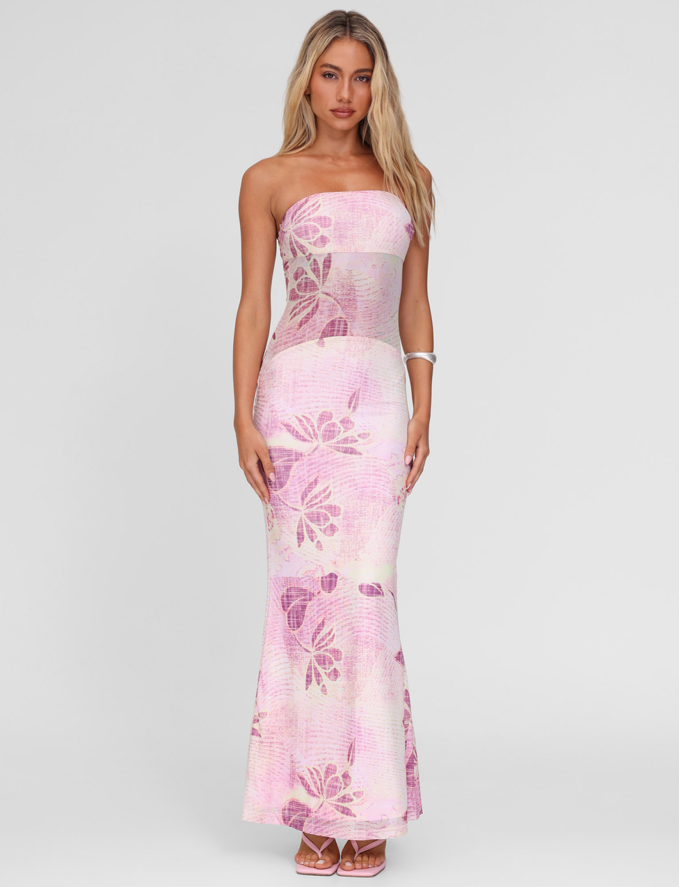 A woman faces forward in the TIGERMIST MORGAN MESH DRESS - GRUNGE FLORAL MAGENTA, a strapless, fitted, floor-length style. With long blonde hair, white sandals, and a bracelet, she stands out against a plain light gray background.