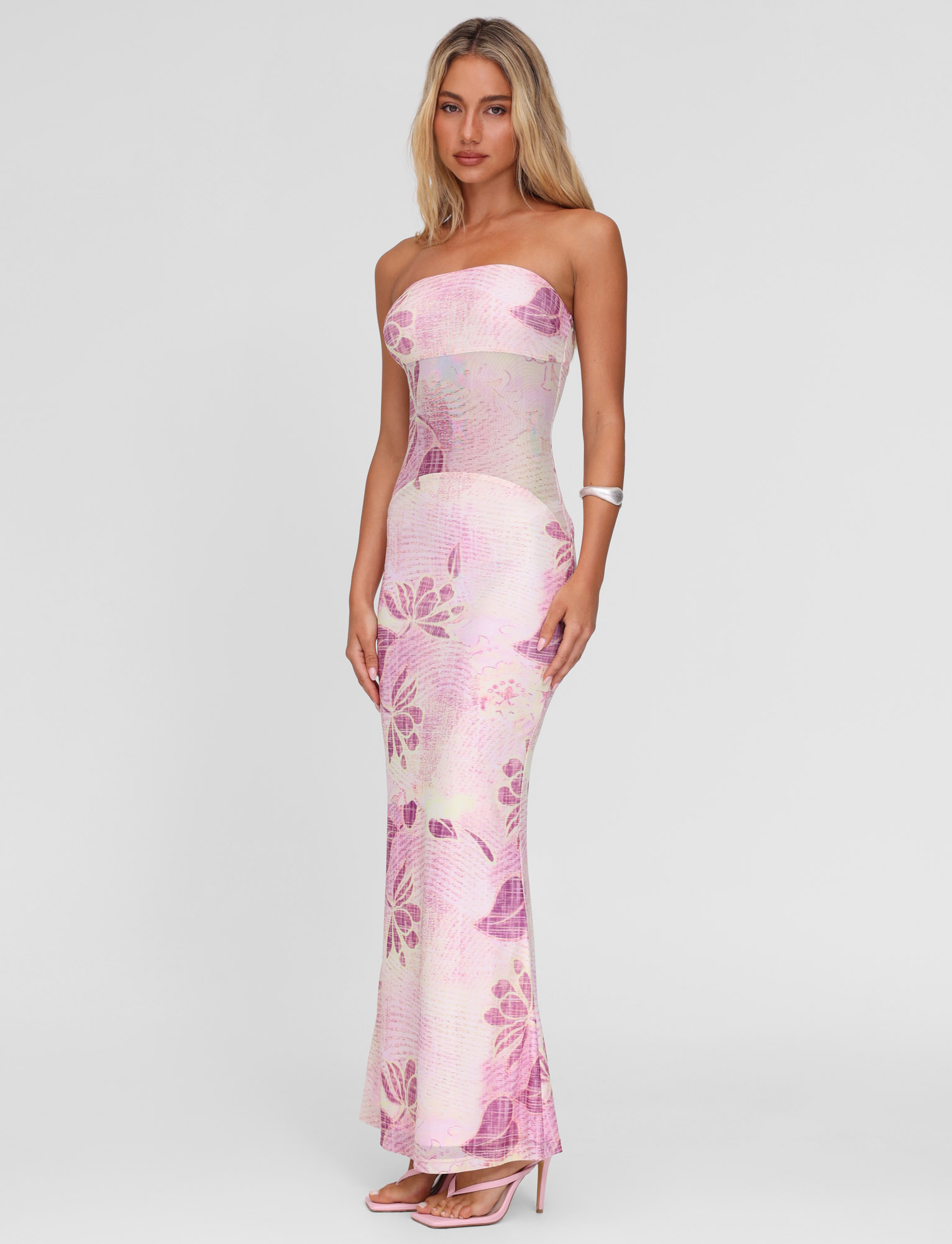 A woman poses in the TIGERMIST MORGAN MESH DRESS - GRUNGE FLORAL MAGENTA, showcasing its strapless neckline, sheer midsection, floor-length silhouette, and pink floral print against a plain white backdrop.