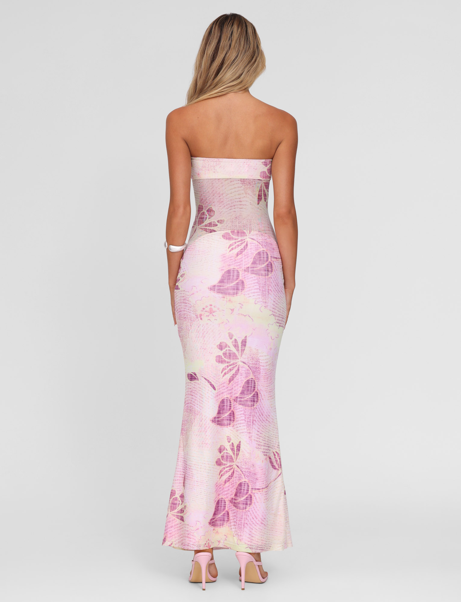 A woman with long blonde hair wears the TIGERMIST MORGAN MESH DRESS - GRUNGE FLORAL MAGENTA, featuring a strapless neckline, styled with pink heels and a white bracelet, shown from the back against a light plain background.