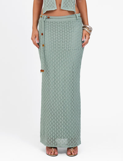 A person standing on a white background wears the stylish COLLINA SKIRT - GREEN by TIGERMIST. The light green crochet maxi skirt with a tie closure and wooden bead details touches the floor. The individual accessorizes with bangles and brown sandals, while their upper body is partially visible.