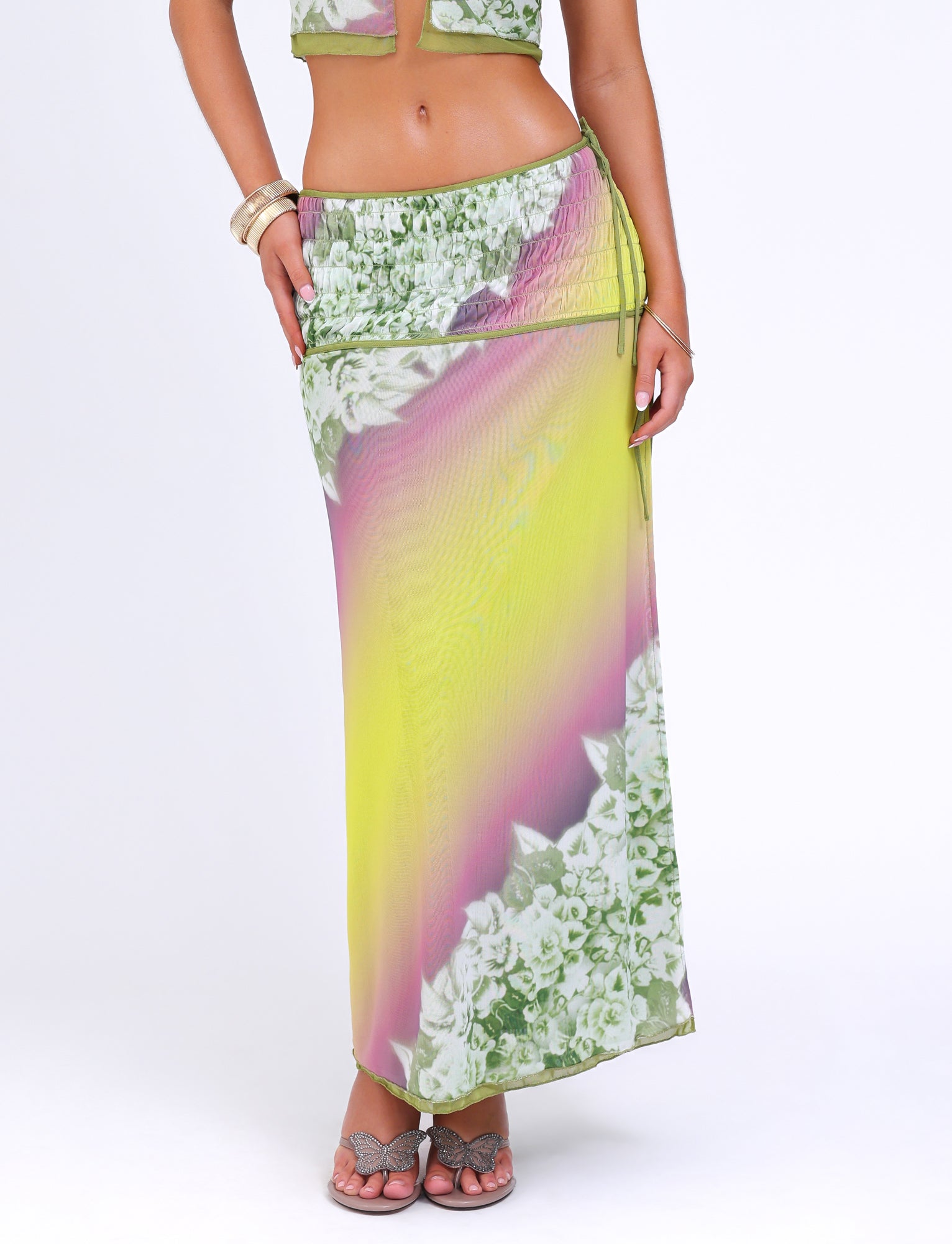 A woman is wearing a vibrant, colorful WAVERLY SKIRT by TIGERMIST, featuring a gradient of green with striped floral patterns and accentuating its low-rise waist. She is also wearing a matching top and sandals, with a bangle bracelet on her wrist against a plain white background.