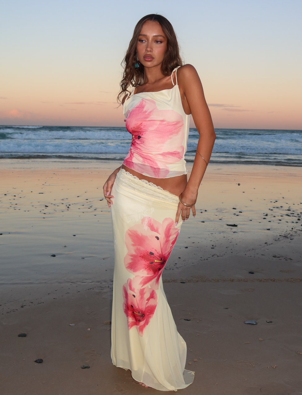 A woman stands on a beach at sunset, wearing the TIGERMIST LILLIAN TOP in yellow. The sea and pastel-toned sky provide a serene backdrop.