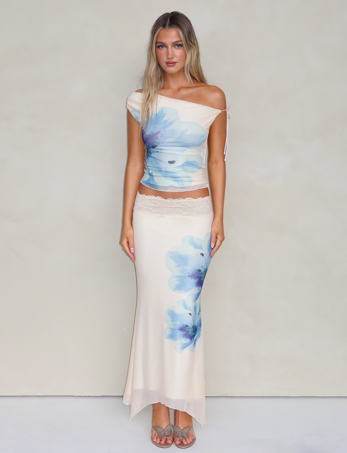 A woman with long, straight blonde hair wears the TIGERMIST LILLIAN SKIRT in white and cream floral, paired with an off-shoulder cream top and sandals, standing against a plain light background.
