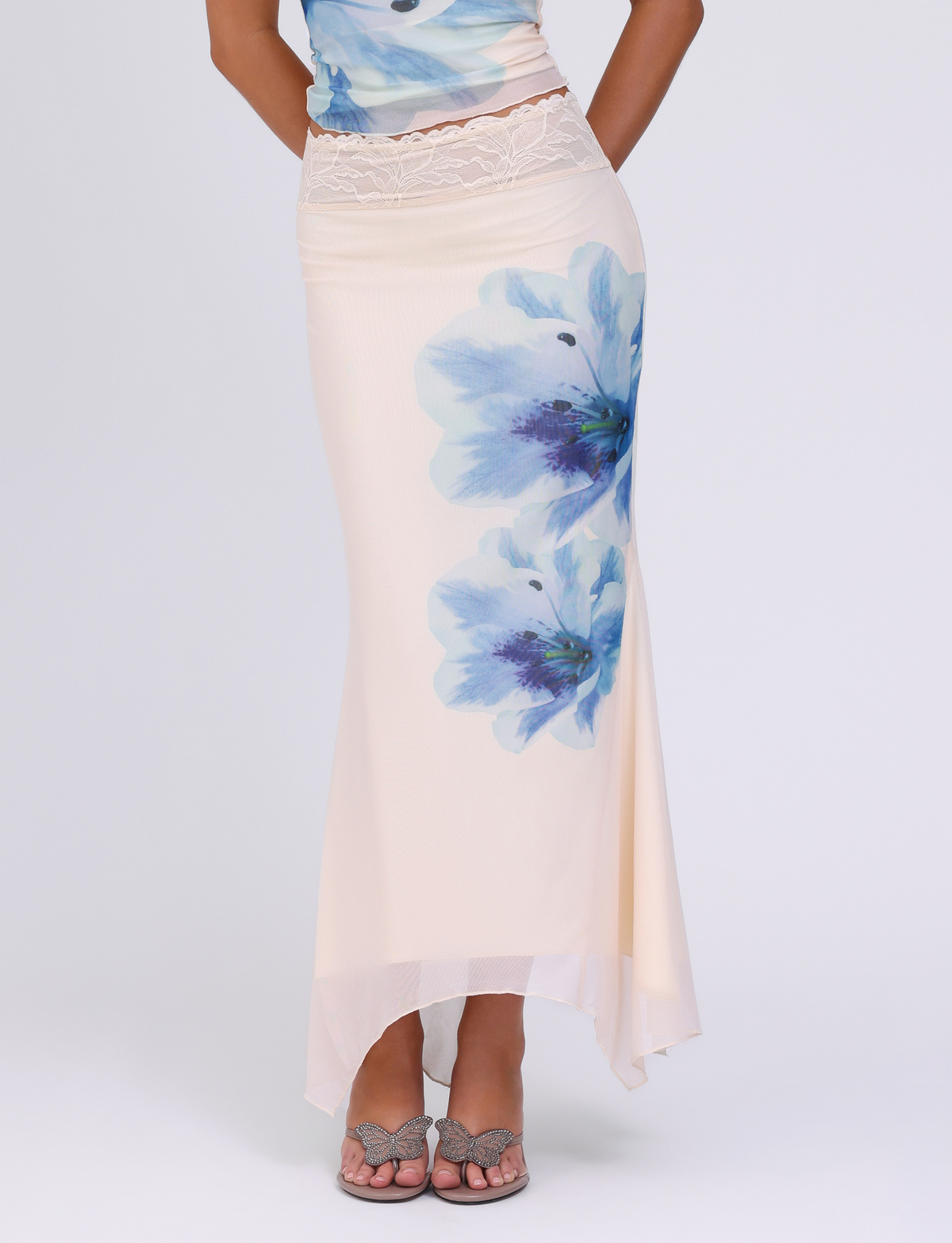 Wearing the TIGERMIST LILLIAN SKIRT in cream with blue floral print and white lace waist detailing, styled maxi length and paired with open-toe heeled sandals. The upper body is partly visible.