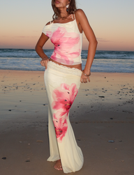 A woman stands barefoot on the beach at sunset, wearing the TIGERMIST LILLIAN SKIRT in yellow. The matching cream top and floral skirt with large pink blooms contrast with the serene ocean and pastel sky backdrop.
