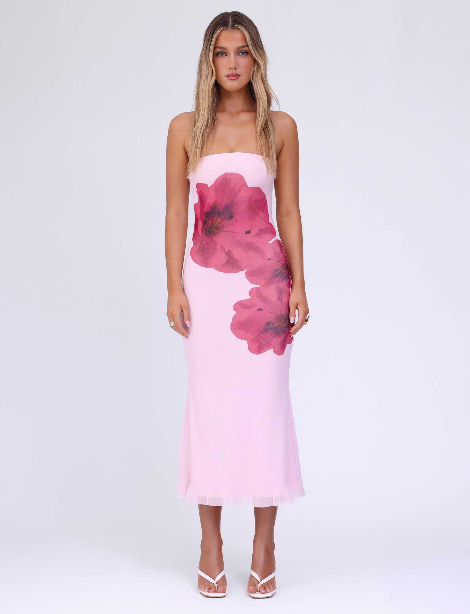 A woman with long blonde hair stands against a plain white background, wearing the strapless LILLIAN DRESS - PINK : BABY PINK by TIGERMIST, adorned with large red floral prints. The midi-length dress pairs perfectly with her white open-toe high-heeled shoes. Her expression is neutral.