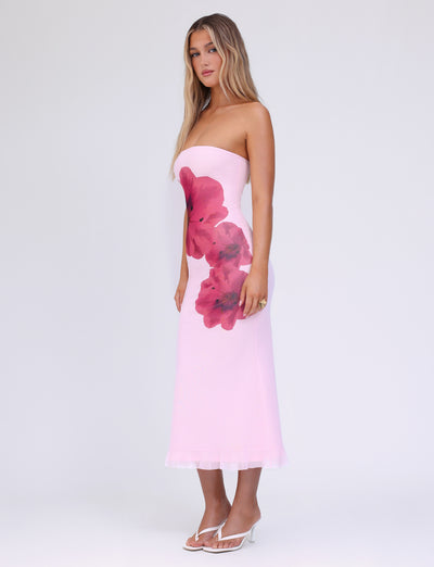 A woman stands against a plain white background wearing the TIGERMIST LILLIAN DRESS in baby pink. The light pink, strapless, floral print dress is midi length. She has long, wavy blond hair and is wearing white heeled sandals. She poses with her body angled slightly to the side.