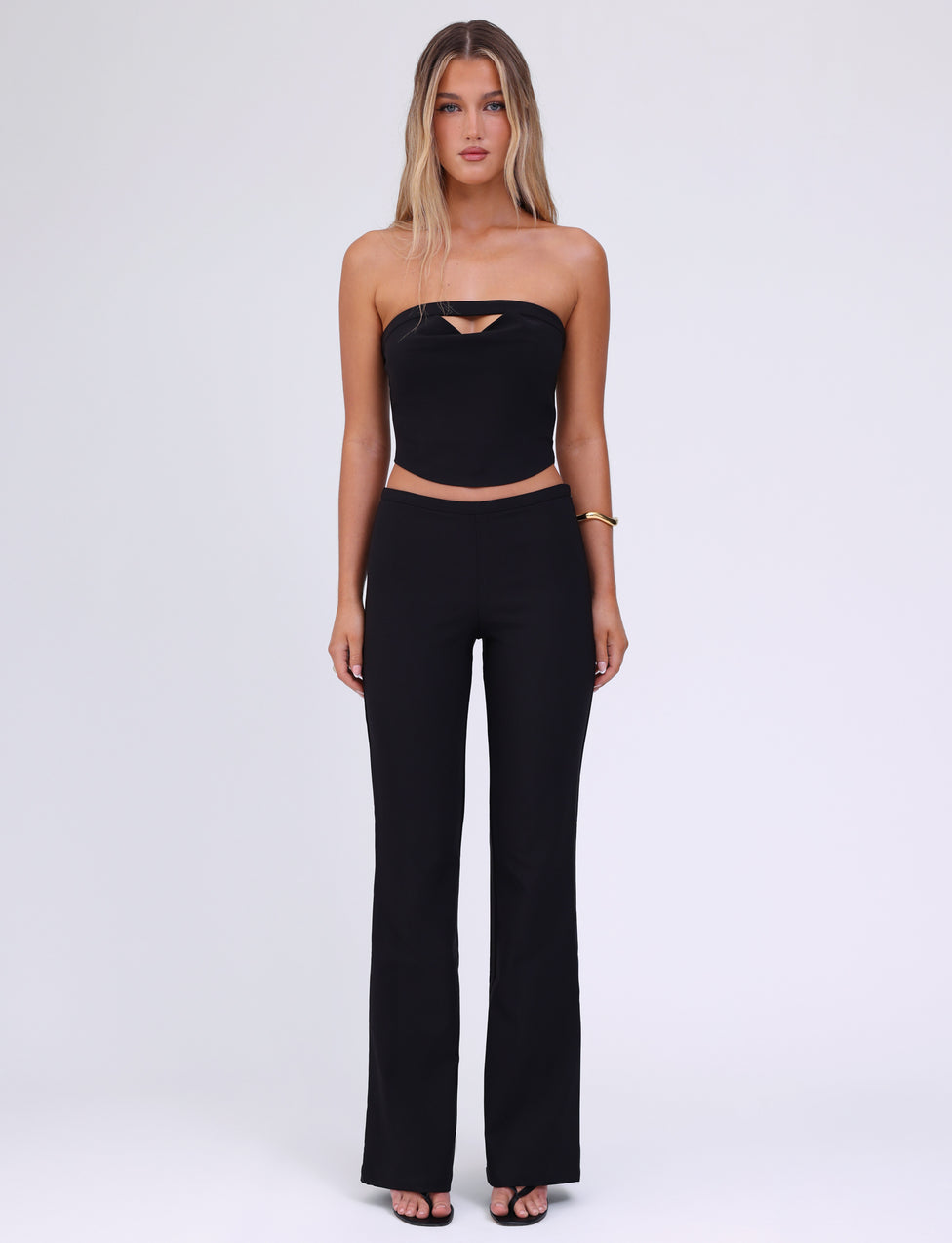 A woman with long, blonde hair stands against a plain white background. She is wearing a black tube top with a small cut-out at the chest, the TIGERMIST AUDRIE PANT in black high-waisted bell-bottom style made from suiting fabrication, and a faux leather belt. She has a gold bracelet on her left wrist and wears black open-toe shoes.
