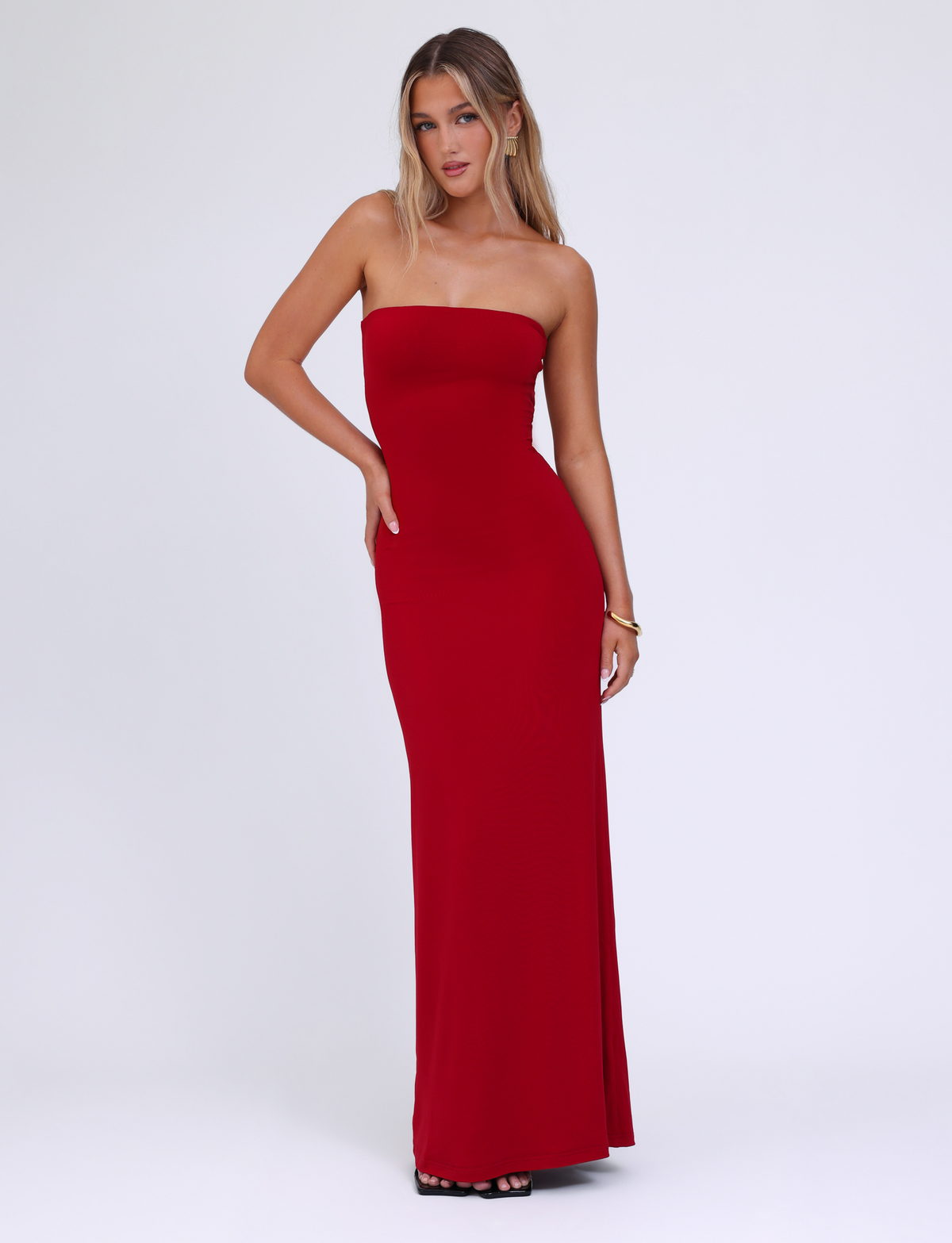 A woman with long blonde hair and a neutral expression poses against a white background, wearing the TIGERMIST DANIELA DRESS - RED. The dress is strapless, form-fitting jersey with a straight neckline. She completes the look with black high heels.
