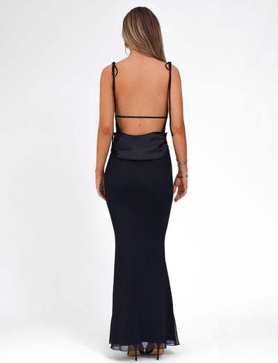 A woman stands with her back to the camera, wearing the TIGERMIST CYRIL DRESS - BLACK. This elegant dress features a low-cut back and thin, adjustable shoulder straps. Her long, straight hair cascades down as she stands in high heels. The background is a plain white studio setting.