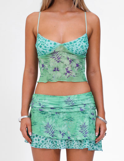 A person is wearing the TIGERMIST SAIGE TOP in green, which features a mixed pattern of leopard spots, palm leaves, and floral prints. The matching ensemble includes an adjustable elastic top that gives it a light, summery appearance. The individual has long blonde hair and is accessorizing with silver bracelets on both wrists.