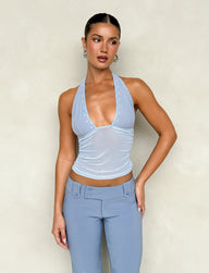 Against a beige backdrop, a person poses confidently, wearing the TIGERMIST BRESILY TOP in the steel blue colorway paired with high-waisted pants. Their sleek hairstyle is complemented by earrings, and their hand rests on their hip.