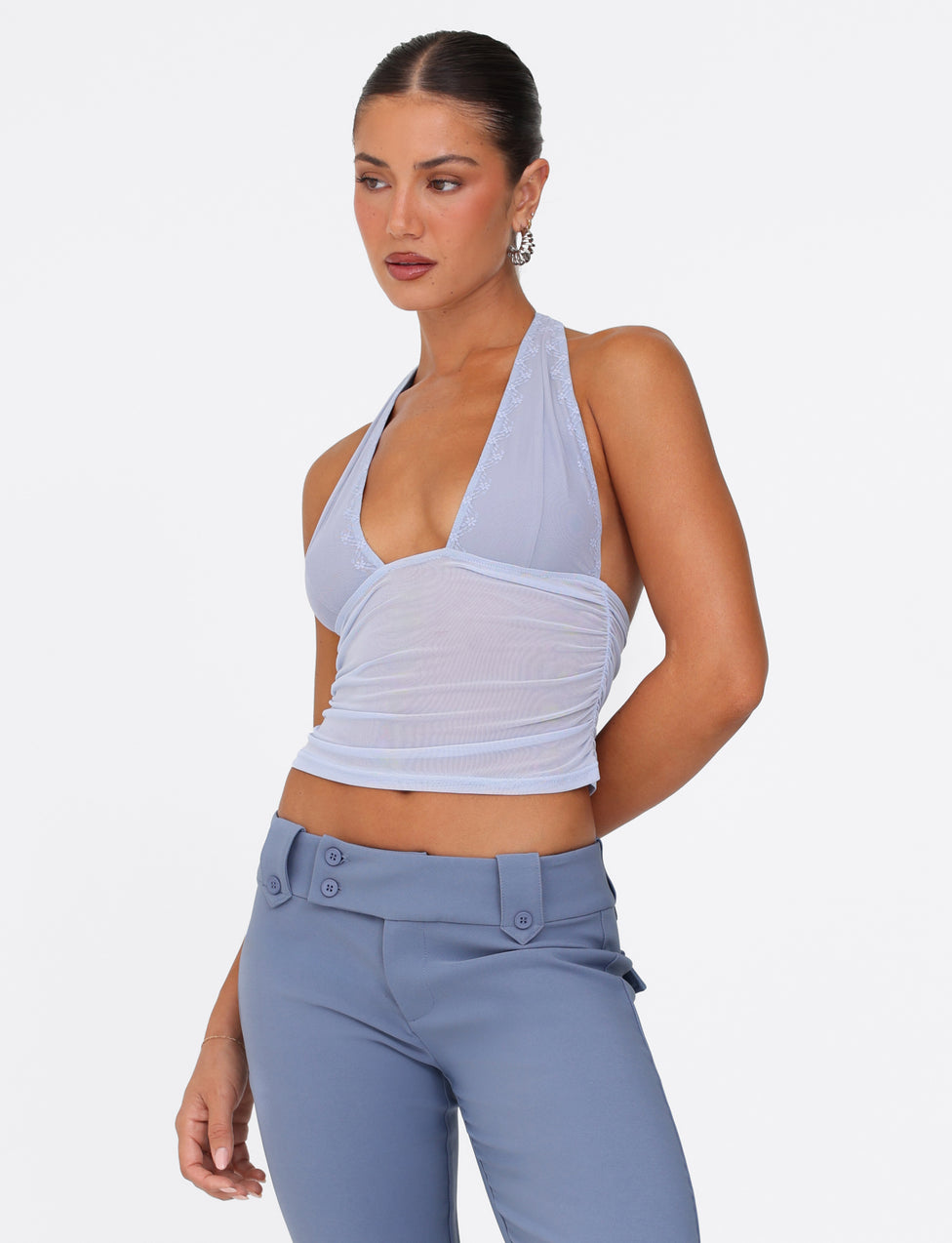 A woman with dark hair tied back is wearing a TIGERMIST BRESILY TOP in steel blue and high-waisted pants against a plain white background. She is looking to the side with a neutral expression.