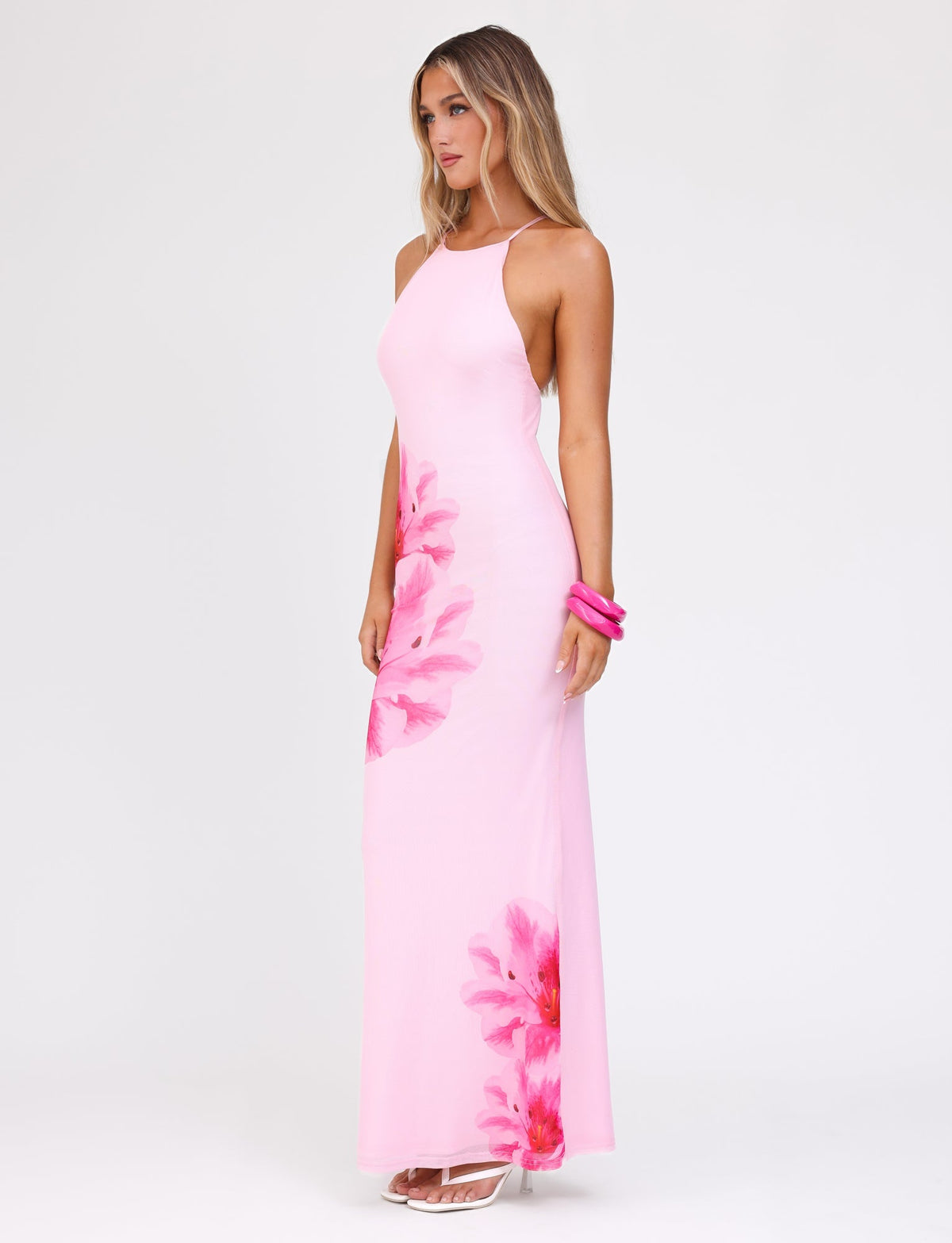 A woman stands against a plain white background, wearing the TIGERMIST KARTER DRESS in baby pink with a maxi length and floral mesh fabrication. She has long, wavy hair, accessorized with pink bangles on her wrist and white high-heeled sandals.