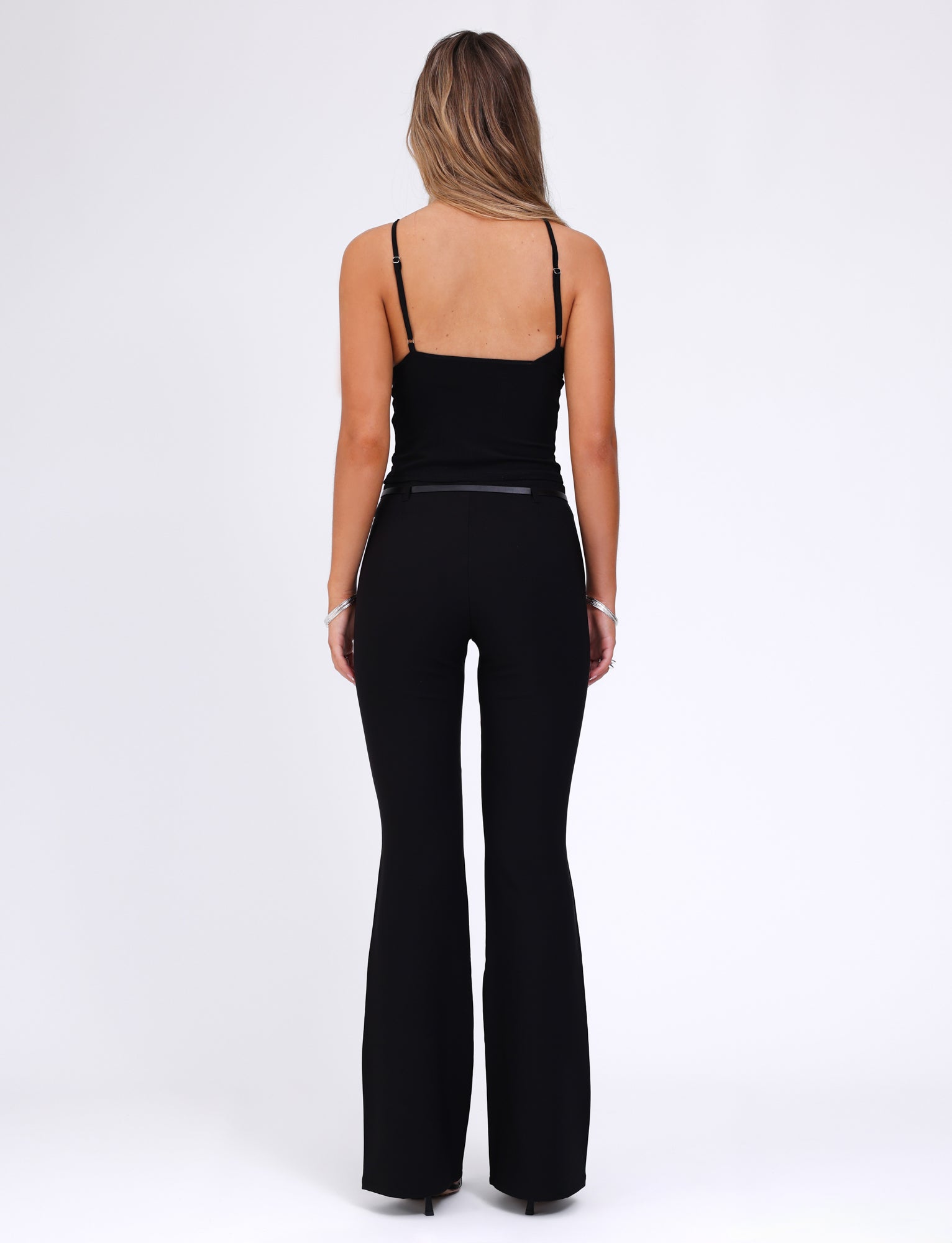 A person with long hair is standing with their back to the camera. They are wearing a black sleeveless top and the TERLA PANT - BLACK from TIGERMIST, featuring a mid-rise waist and a flared silhouette. The background is plain white.