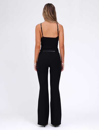 A person with long hair is standing with their back to the camera. They are wearing a black sleeveless top and the TERLA PANT - BLACK from TIGERMIST, featuring a mid-rise waist and a flared silhouette. The background is plain white.