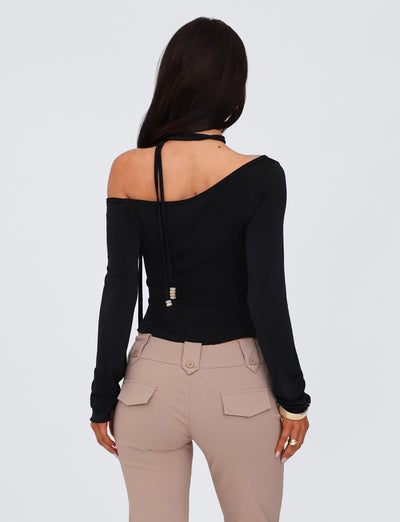 A person with long dark hair stands facing away from the camera, wearing a TIGERMIST MIETTA TOP - BLACK, which features a multi-tie neck and high stretch fabric. Light beige pants with flap pockets and buttons complete the look against a plain, light-colored background.