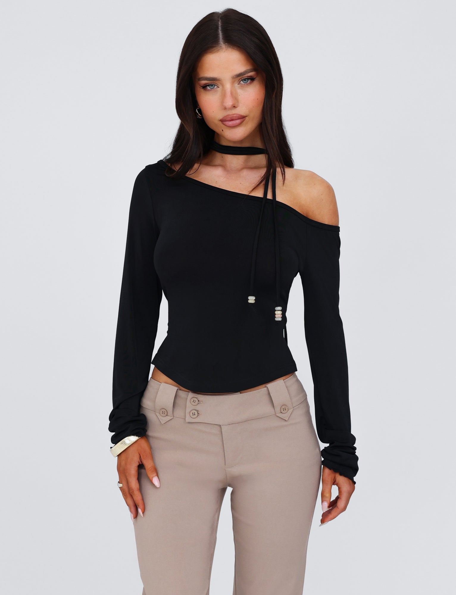A woman with long brown hair stands facing the camera. She is wearing the TIGERMIST MIETTA TOP in Black, featuring a choker-style neckline with hanging strings and a multi-tie neck. The high-stretch fabric of her off-the-shoulder top complements her long-sleeved look paired with beige high-waisted pants. Her hands are relaxed by her sides.
