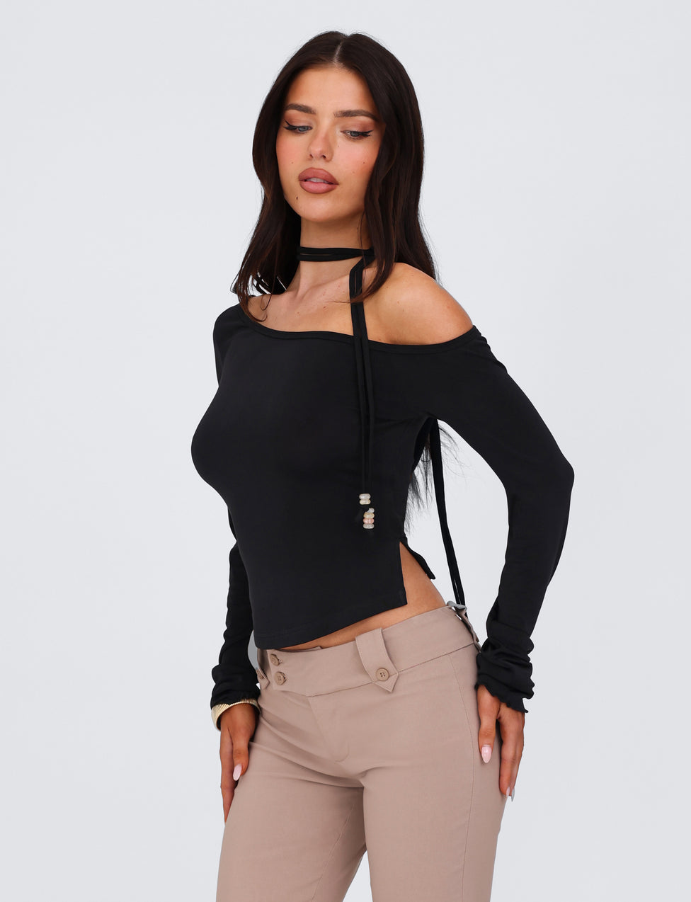 A woman with long dark hair is wearing a Black Mietta Top from TIGERMIST, an off-the-shoulder long-sleeve design made of high stretch fabric featuring a multi-tie neck, paired with beige pants. She is looking down and slightly to the side, her hands resting on her thighs. The background is plain and light-colored.