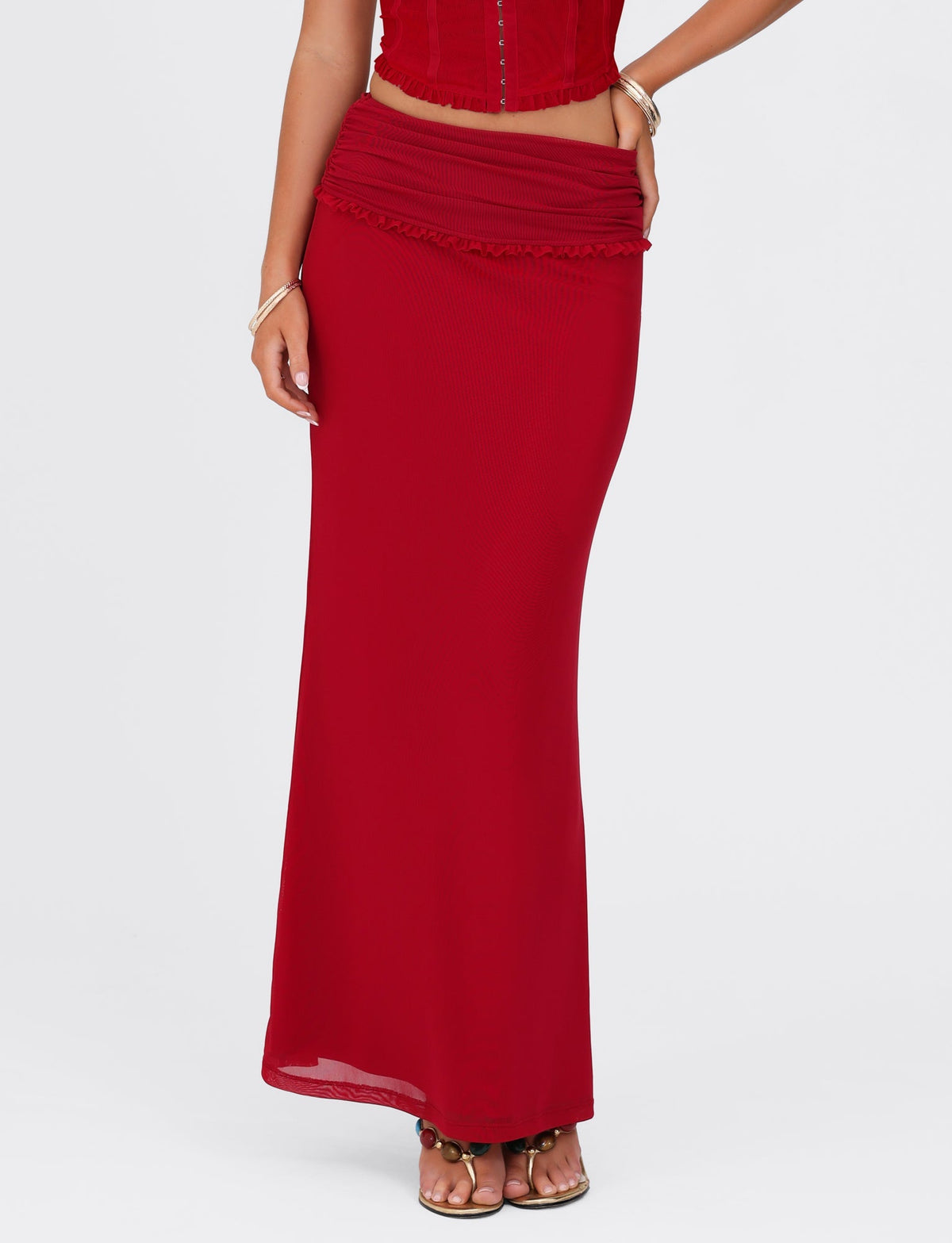 A person is wearing the TIGERMIST GINNY MAXI SKIRT - RED : CHERRY RED, which extends to the floor, paired with a matching red top. Their arms are relaxed by their sides, showcasing bracelets on one wrist. The outfit exudes style and elegance, with the maxi-length skirt featuring a slight ruffle at the top.