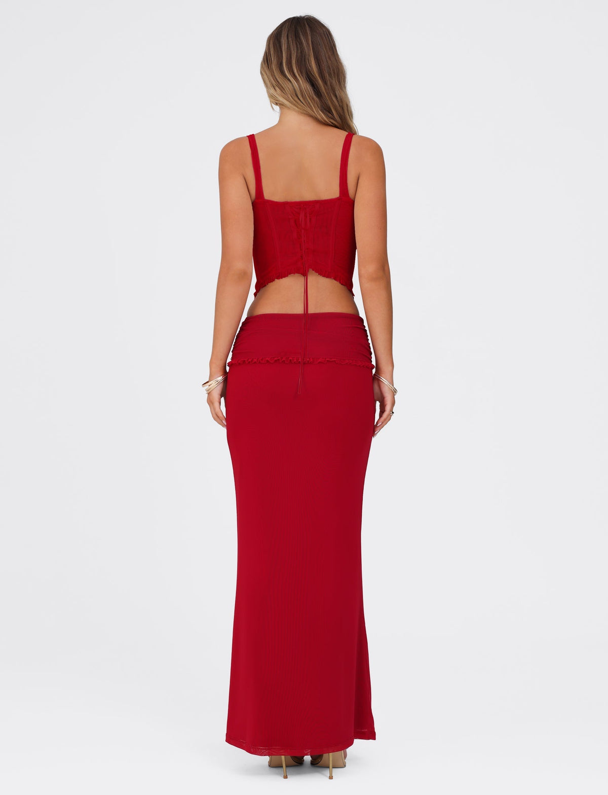 A woman with long hair is seen from the back, wearing a red, sleeveless cropped top with a bow at the back and a matching high-waisted GINNY MAXI SKIRT - RED in Cherry Red by TIGERMIST. The outfit is made of stretchy fabric, and she stands against a plain white background.