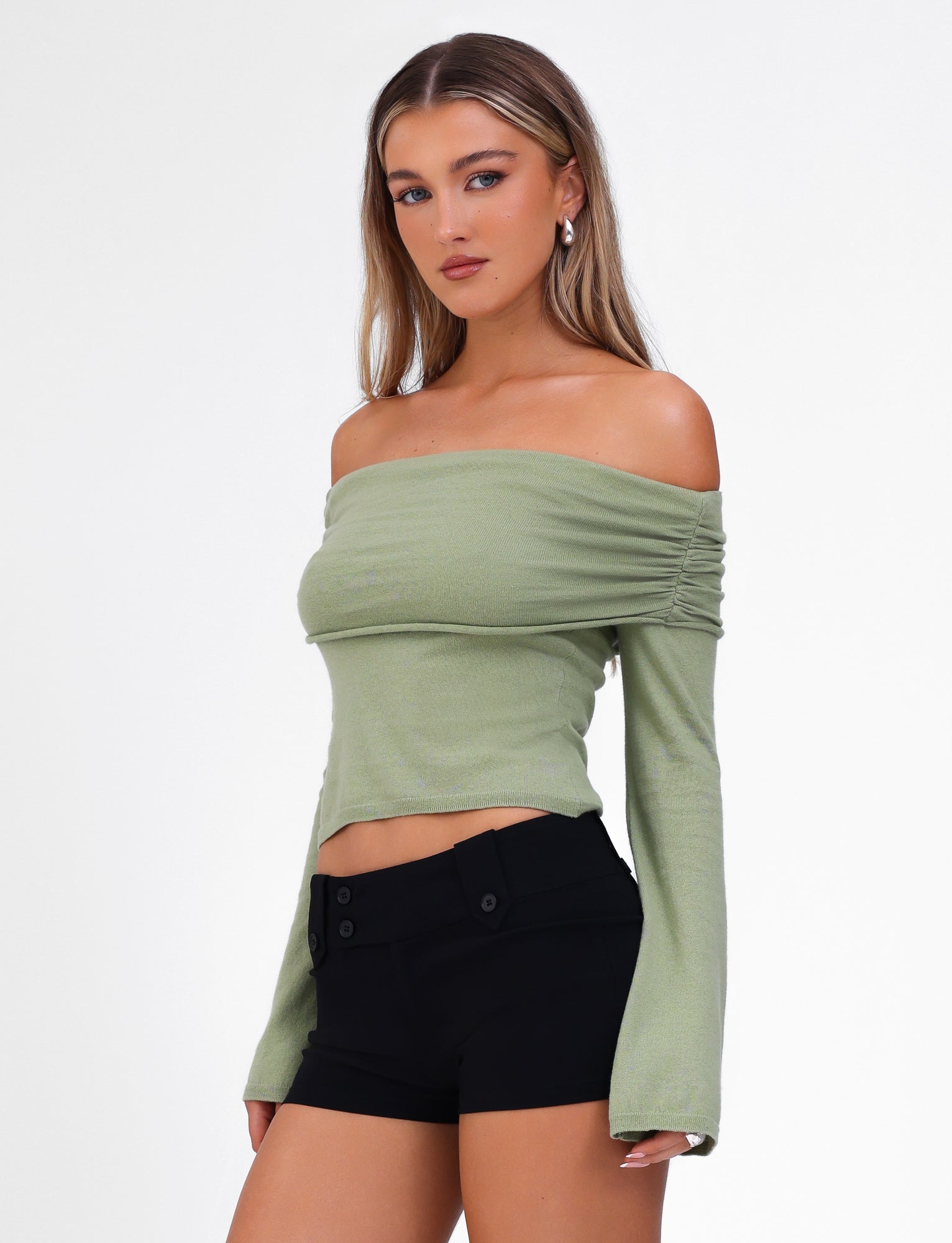 A woman with long, straight hair wears the DIEDRE JUMPER - GREEN from TIGERMIST, a light green off-the-shoulder top with long sleeves paired with black high-waisted shorts. The off-shoulder neckline highlights her collarbones as she stands confidently against a plain white background. Her minimal makeup accentuates her natural beauty.