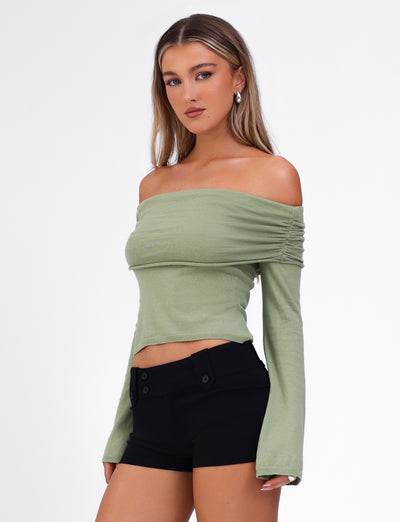 A woman with long, straight hair wears the DIEDRE JUMPER - GREEN from TIGERMIST, a light green off-the-shoulder top with long sleeves paired with black high-waisted shorts. The off-shoulder neckline highlights her collarbones as she stands confidently against a plain white background. Her minimal makeup accentuates her natural beauty.
