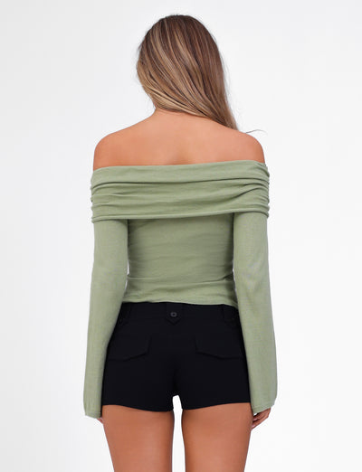 A woman with long, wavy hair stands with her back to the camera, showcasing the DIEDRE JUMPER - GREEN from TIGERMIST. The off-the-shoulder, long-sleeved moss green top is made of high stretch fabric and paired with high-waisted black shorts. The background is plain white.