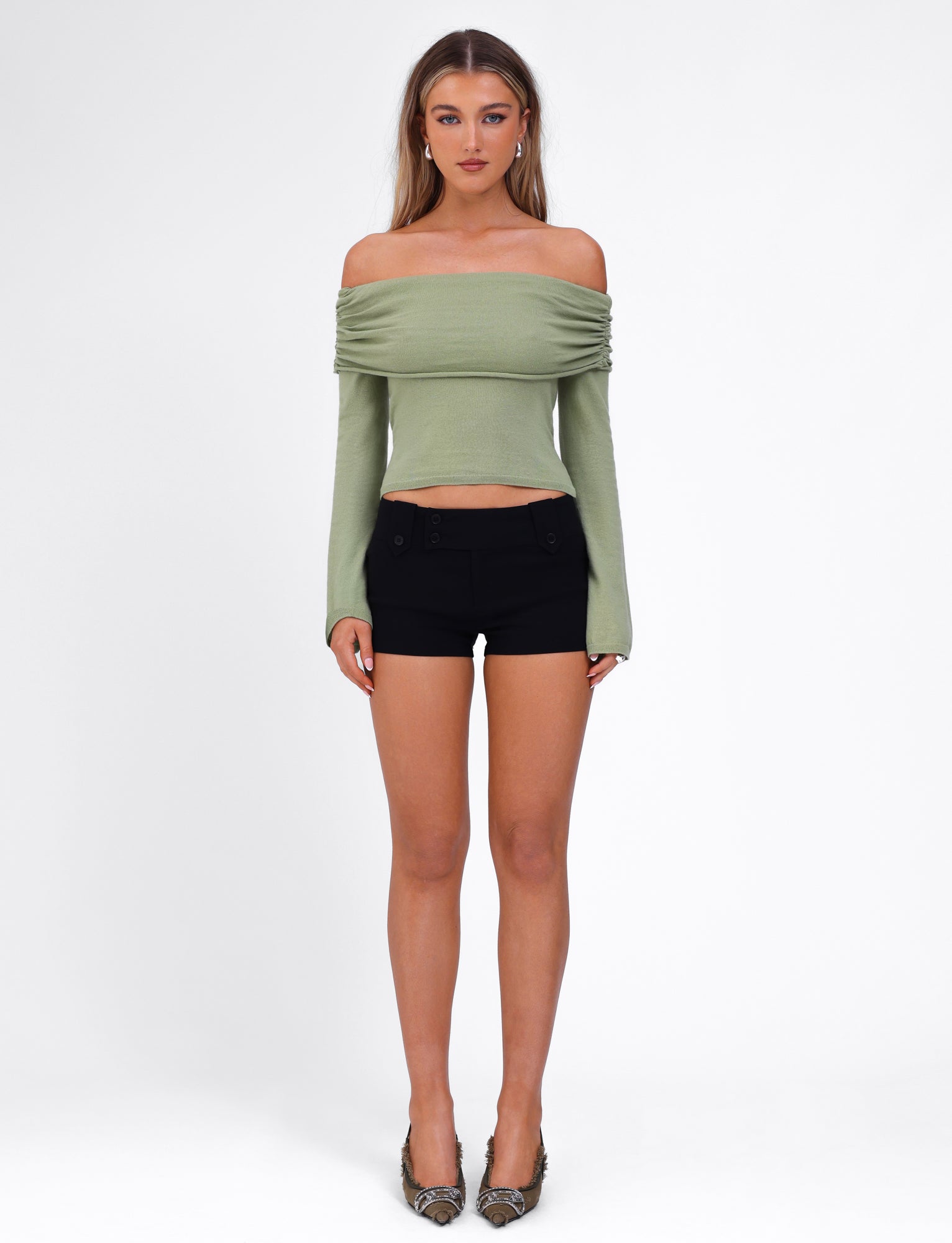 A woman stands against a plain white background wearing the DIEDRE JUMPER - GREEN from TIGERMIST, an off-the-shoulder light green long-sleeve top made of high-stretch fabric, paired with black shorts. She has long, straight hair and is wearing high-heeled shoes. Her arms are relaxed at her sides as she looks straight at the camera.