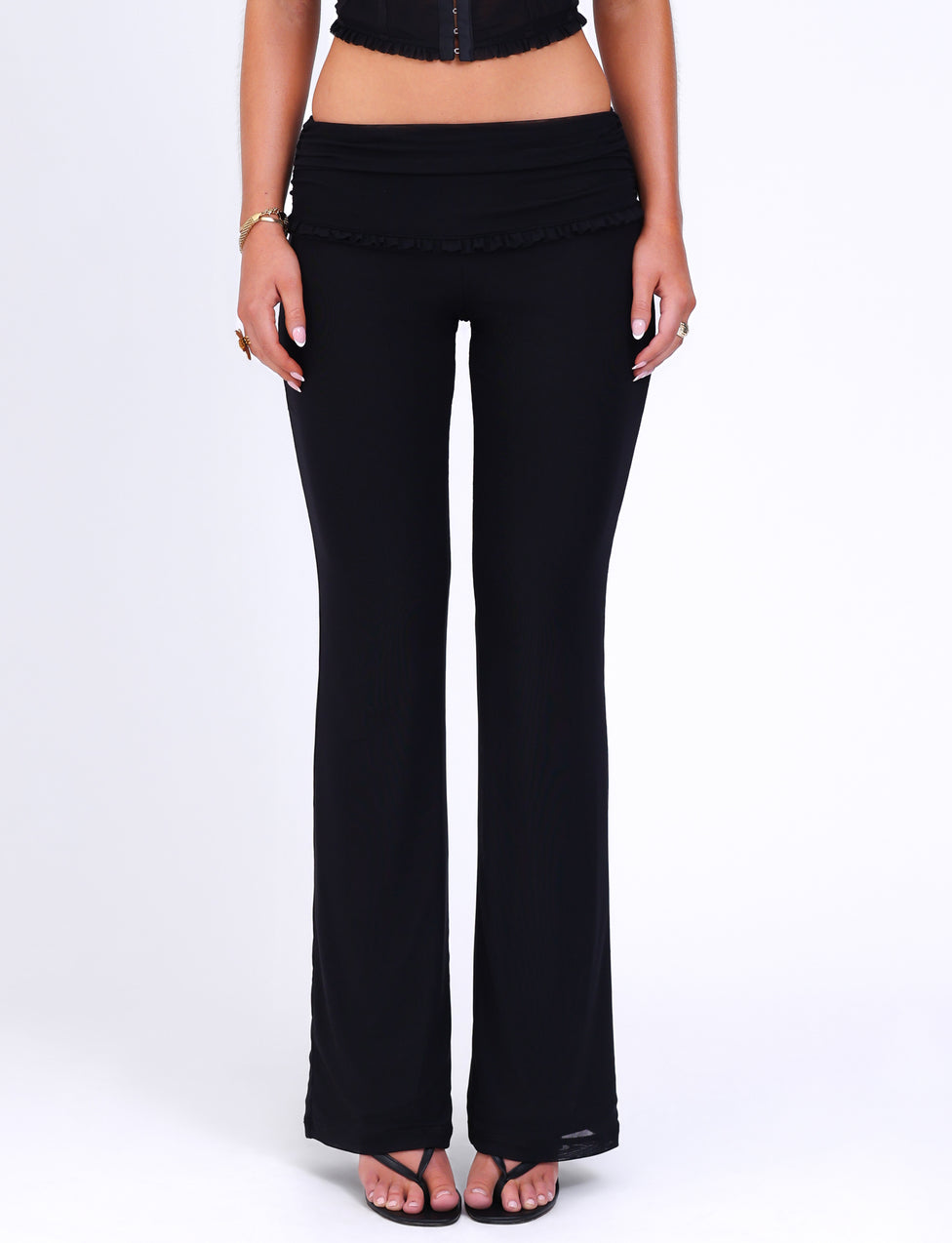 A person is adorned in TIGERMIST's GINNY PANT - BLACK, complemented by black open-toe sandals, standing against a white backdrop. The individual, also wearing a cropped black top, is photographed from the waist down to emphasize the chic flared silhouette.