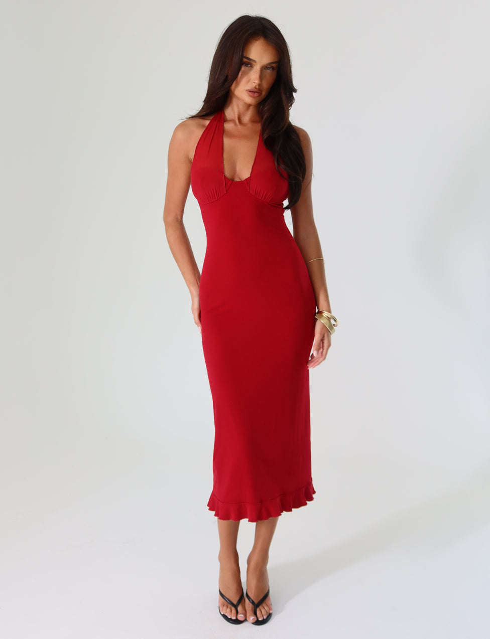 A woman wearing the TIGERMIST GRANGER MIDI DRESS in cherry red—featuring a fitted halter-neck design with ruffle details—poses against a plain background. The dress's flared hem is stylishly paired with black high-heeled sandals and gold bracelets on her wrist.