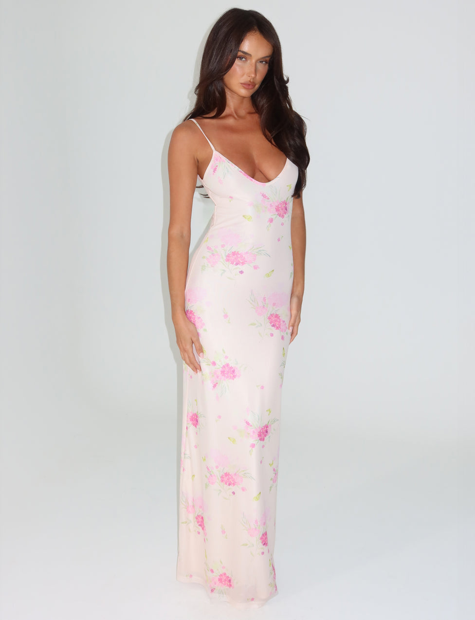 A woman wearing the OSIRIS DRESS from TIGERMIST, adorned in a watercolour floral print with adjustable shoulder straps, stands against a plain background. Her long dark hair flows naturally as she gazes directly at the camera with a neutral expression.