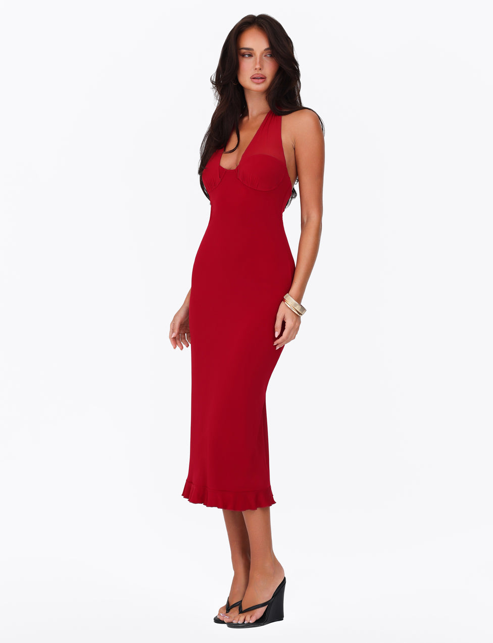 A woman poses in the TIGERMIST GRANGER MIDI DRESS in Cherry Red, showcasing its figure-hugging fit, ruffle detail, and halter neckline. She complements her look with black wedge sandals and gracefully drapes her long, dark hair over one shoulder against a plain white background.