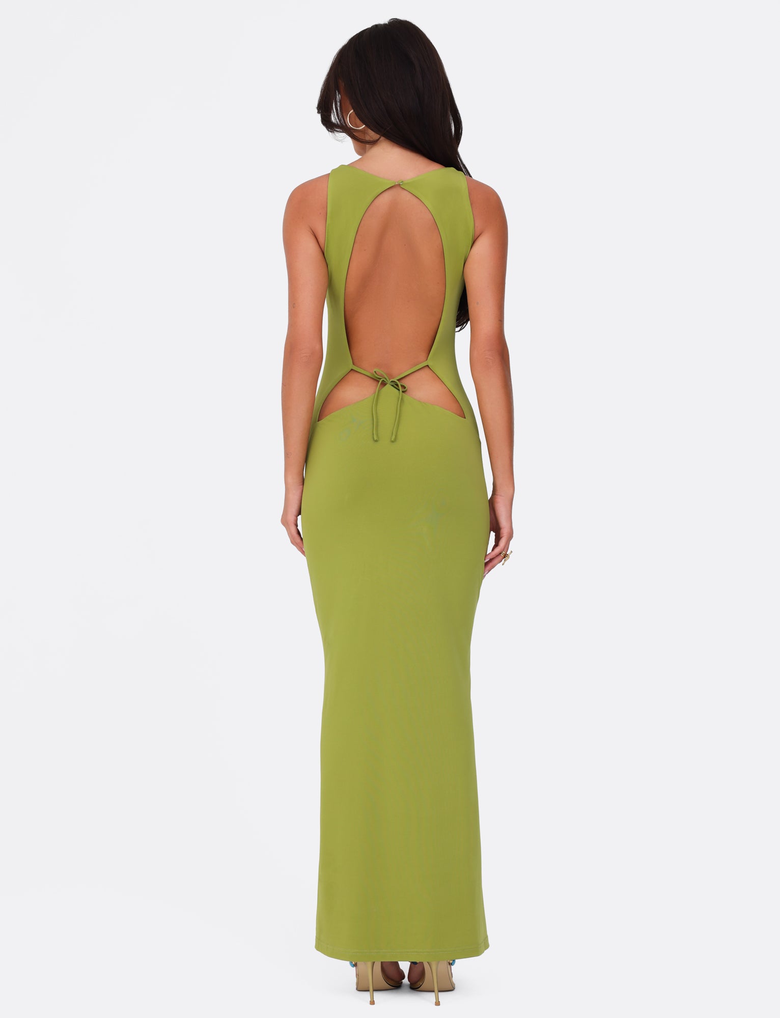 A woman in the SONNET MAXI DRESS - GREEN by TIGERMIST stands facing away. She has long hair and is wearing high heels. The dress features a sweetheart neckline, tie detail, halter neck, and maxi-length skirt against a plain white background.