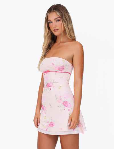 A person with long, wavy hair poses against a plain white background wearing the CARSYN MINI DRESS in pink with a Water Colour Floral print by TIGERMIST. The strapless dress features an asymmetrical hemline that adds a touch of elegance, as one hand rests by their side and the other is slightly bent.