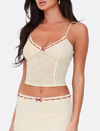 A woman is wearing the FENNEL TOP in pale yellow from TIGERMIST, featuring red bow accents and delicate lace trim. This ensemble includes a cami top and skirt in a flattering bodycon fit. Standing against a plain white background, she exudes elegance and charm.