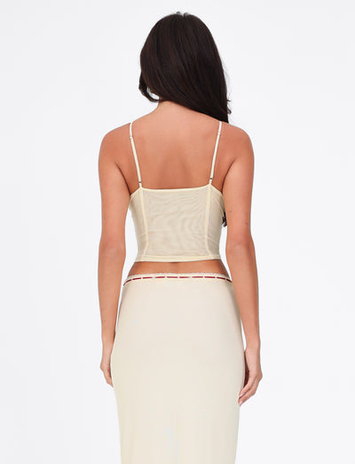 A person with long dark hair is shown from the back, wearing the FENNEL TOP in pale yellow by TIGERMIST and a beige skirt with red lace trim against a plain white background.