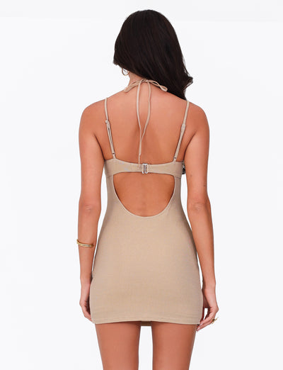 A person stands with their back to the camera wearing the ELLISON MINI DRESS in gold by TIGERMIST. The halter-neck, backless dress has a bodycon fit and features a back cutout with thin straps. Their long hair flows freely, and they wear a bracelet on their right wrist.