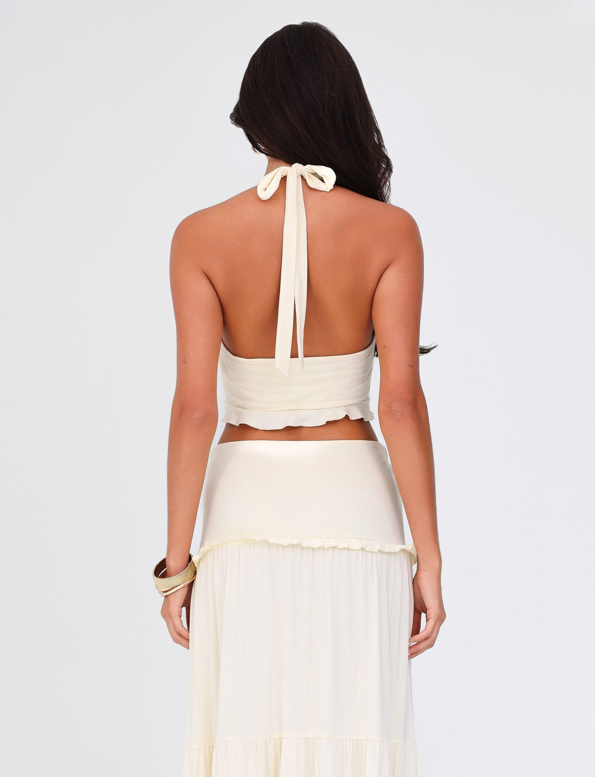 A woman with long dark hair is seen from behind wearing the Tigermist Katya Halter Top - White : Cream and a matching ruffled skirt. The cropped length highlights her waist, and she has a bracelet on her left wrist, set against a plain background.