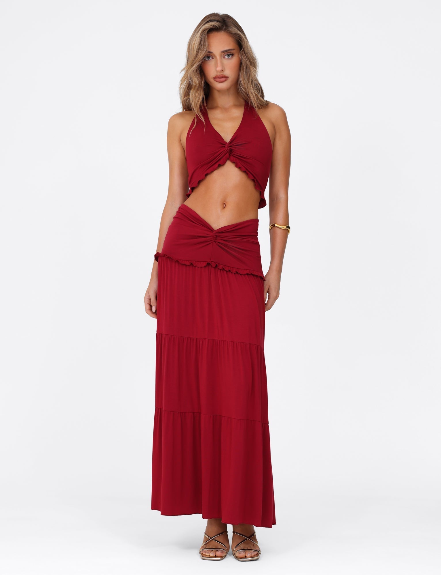 A woman poses against a white background in the KATYA MAXI SKIRT from TIGERMIST, a high-stretch fabric skirt with a wide waistband, paired with a red halter-neck crop top. She accessorizes with strappy sandals and a gold bracelet, her hair styled in loose waves.