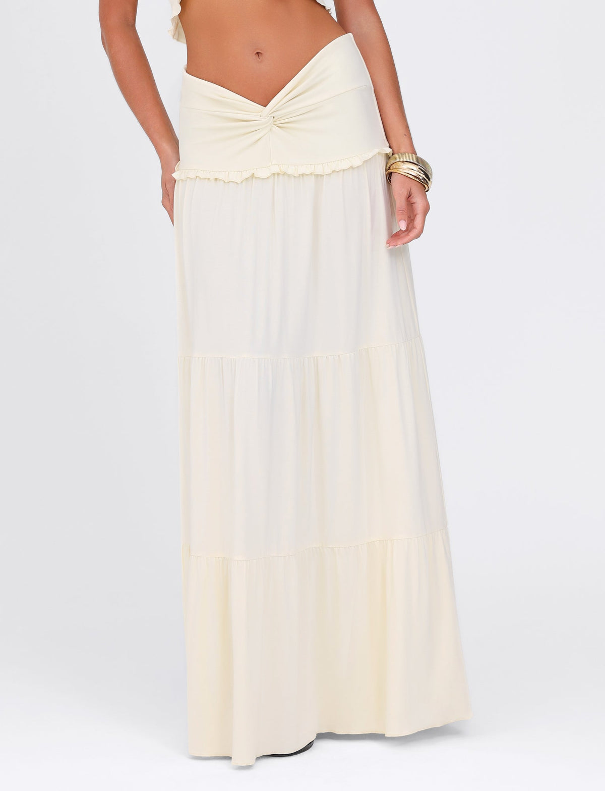 A person wearing the TIGERMIST KATYA MAXI SKIRT in cream, featuring tiered panels and a knotted waistband, stands against a plain white background. Their left wrist is adorned with gold bangles, completing the effortlessly elegant look in buttery jersey.