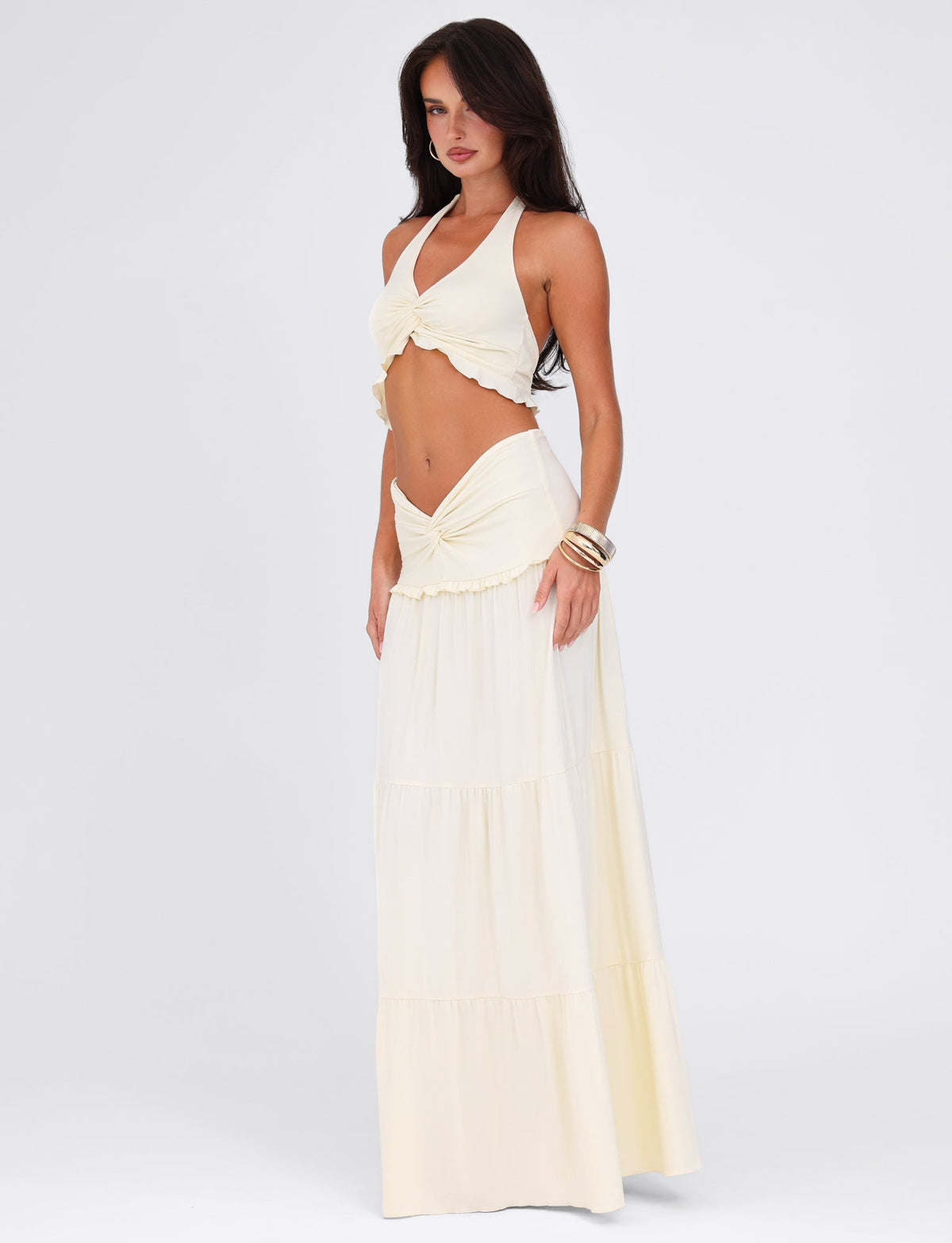 A model poses in a flowing, off-white halter top with tiered panels and the KATYA MAXI SKIRT - WHITE : CREAM by TIGERMIST. The outfit, crafted from buttery jersey, features ruffled details and a high waist. Large bangles adorn one arm as they stand against a plain white background.