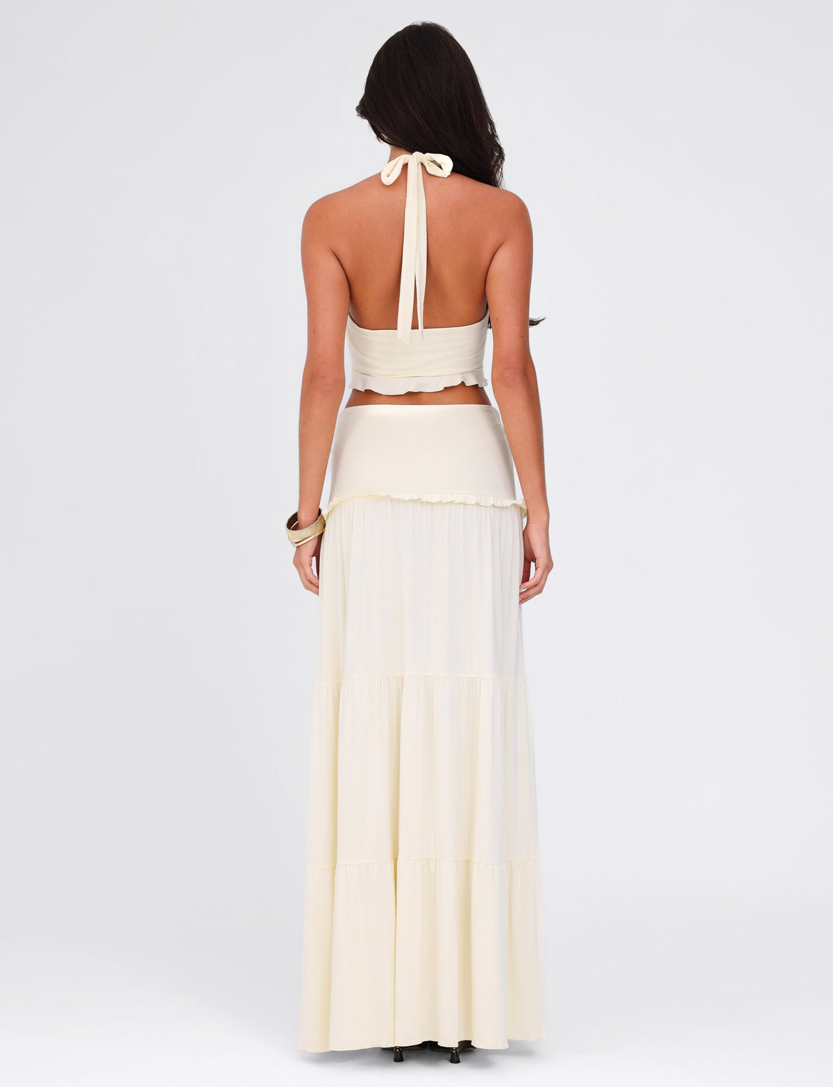 A woman with long dark hair wears a beige, strapless halter top paired with the TIGERMIST KATYA MAXI SKIRT in white and cream. She stands with her back to the camera against a plain white background.