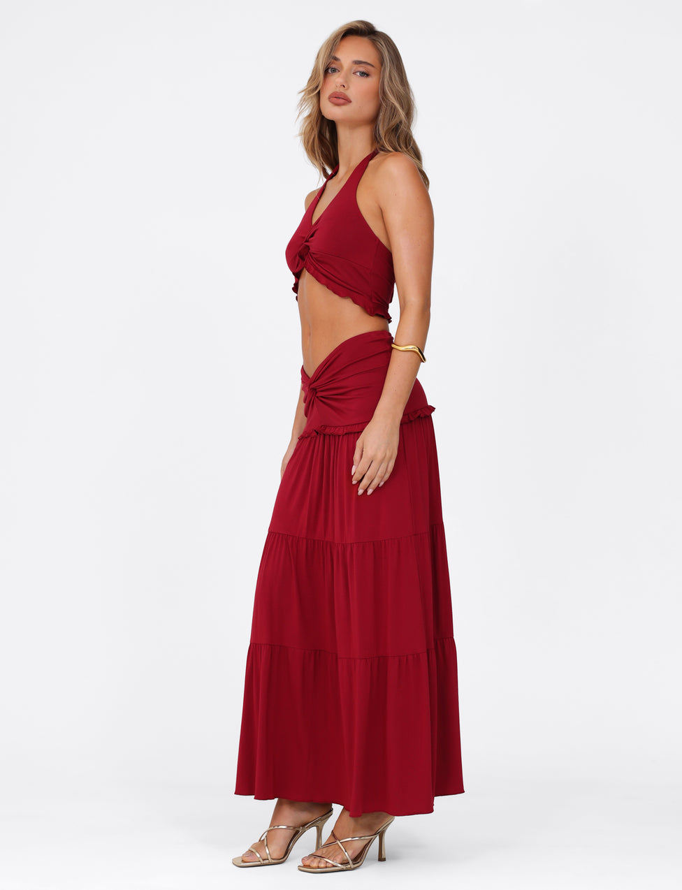 A confident woman poses in TIGERMIST's KATYA MAXI SKIRT - RED and a matching sleeveless crop top. Posing in profile with her left hand on her hip, she dons gold heels, a bracelet, and lets her wavy hair cascade elegantly down.