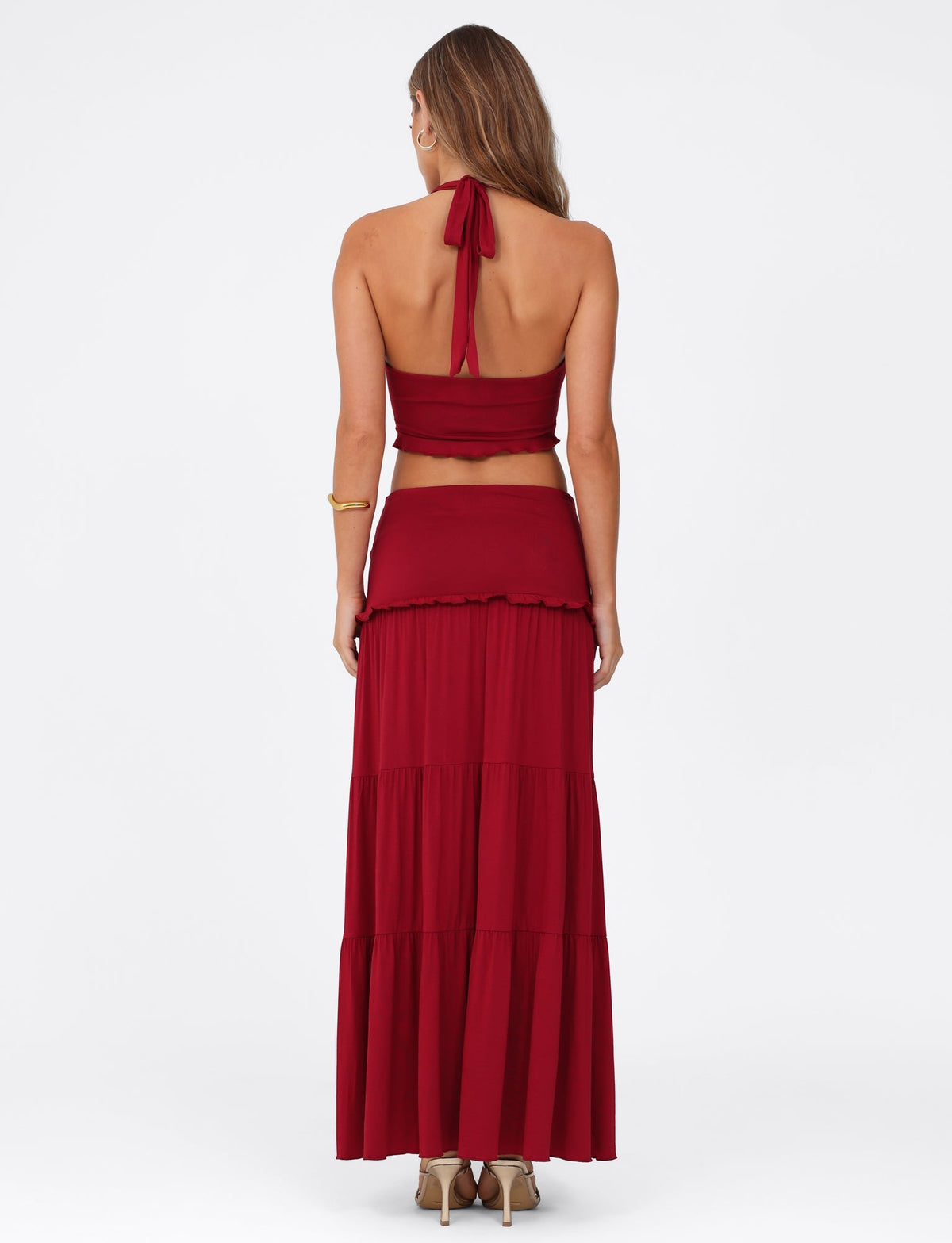 A woman showcases the TIGERMIST KATYA MAXI SKIRT - RED with its wide waistband and tiered design. Her flowing hair pairs beautifully with high-heeled sandals, all captured against a plain white background.