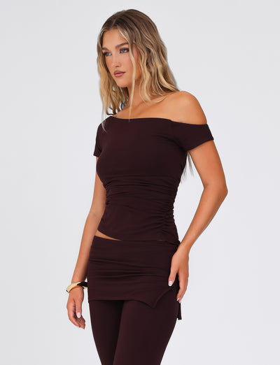 A woman with long hair wears the TIGERMIST LEILANI TOP in chocolate, paired with a matching skirt. She stands against a plain, light-colored background, gazing to the side. Her ensemble boasts gathered details and an asymmetrical neckline, complemented by a bracelet on her right wrist.