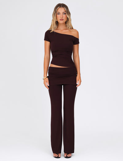 A person wearing a TIGERMIST off-shoulder dark brown top and the matching LEILANI PANT - BROWN: CHOCOLATE stands against a plain white background. They have light brown hair, wear black heels, and accessorize with a bracelet on one wrist.