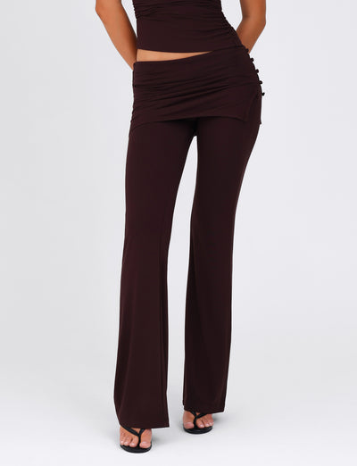 A person is wearing the LEILANI PANT - BROWN : CHOCOLATE by TIGERMIST, featuring a layered waistband and complemented by a matching top. Against a plain white background, they confidently showcase the stylish high stretch fabric, paired with black open-toe shoes and their hands resting on their hips.