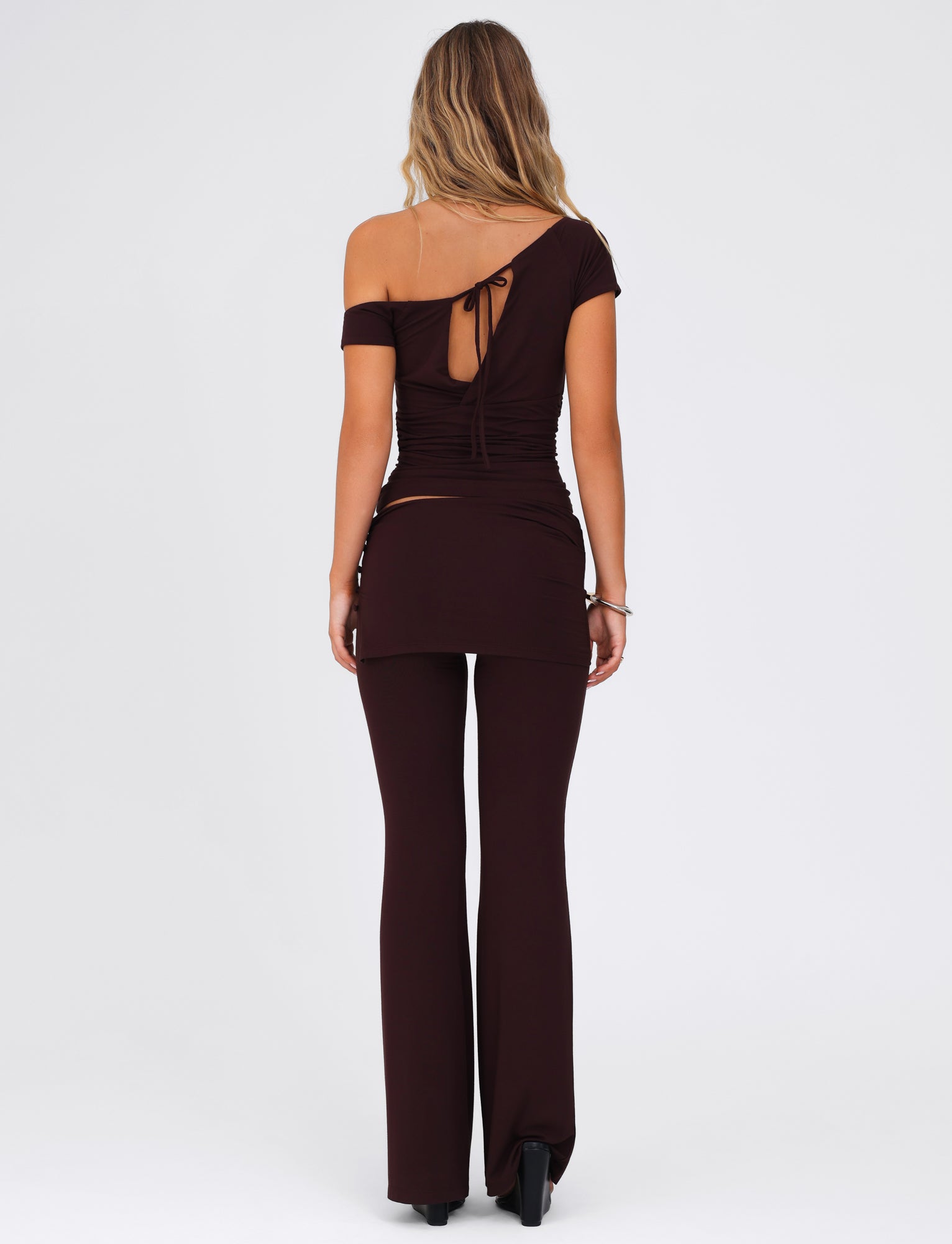 A woman stands with her back turned, wearing the TIGERMIST LEILANI PANT in a chocolate brown hue, accompanied by a chic off-shoulder top featuring a tie at the back and made from high-stretch fabric. Her long, wavy hair flows over the coordinated ensemble against a simple, light background.